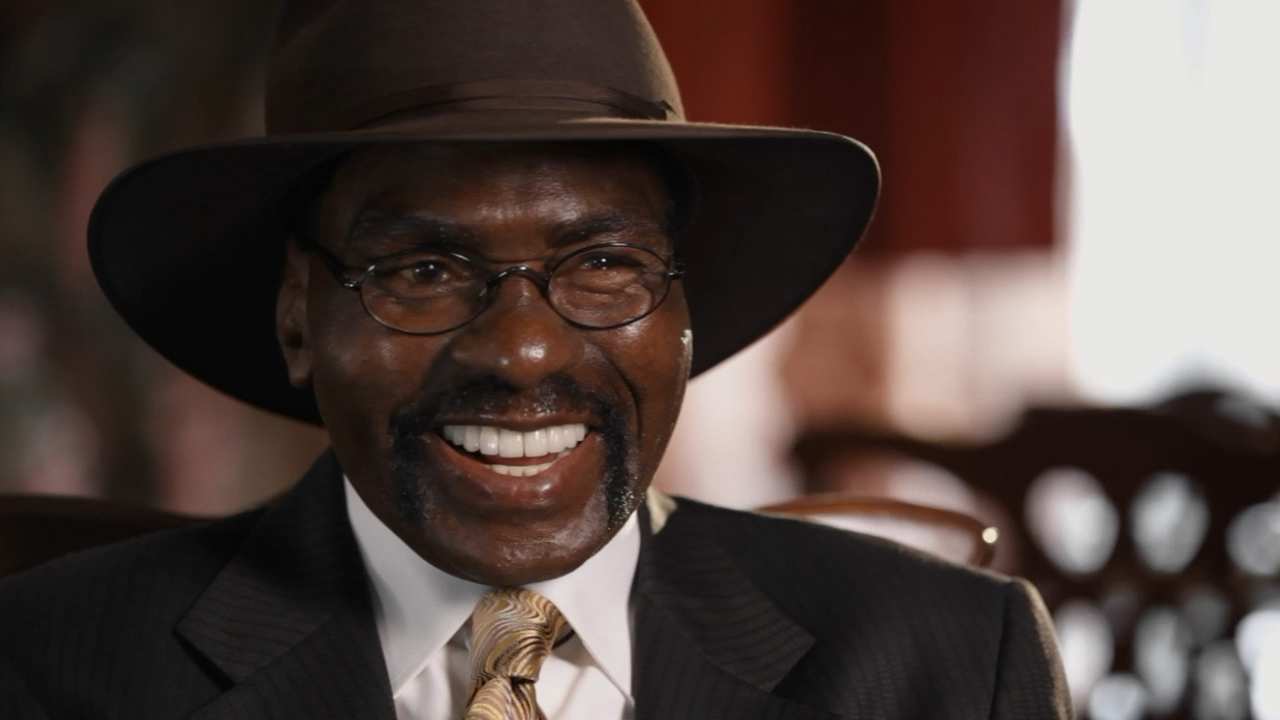 a man with glasses and a hat sitting talking