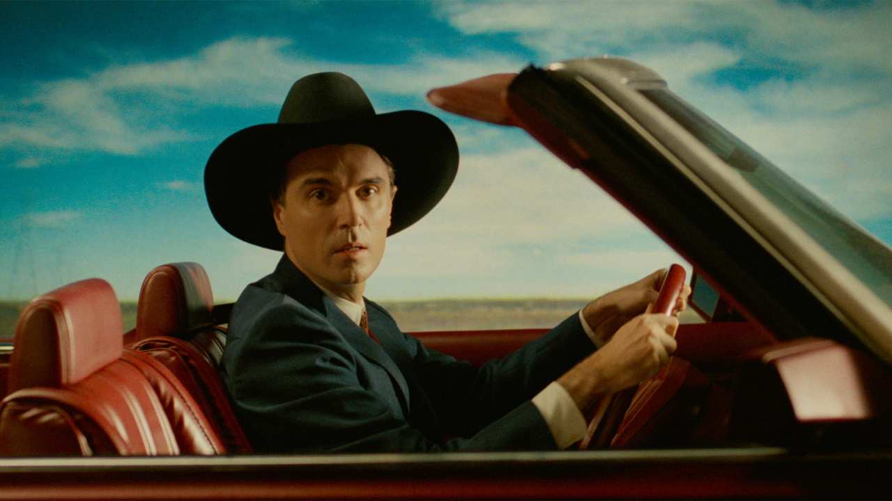 David Byrne in a cowboy hat driving a car
