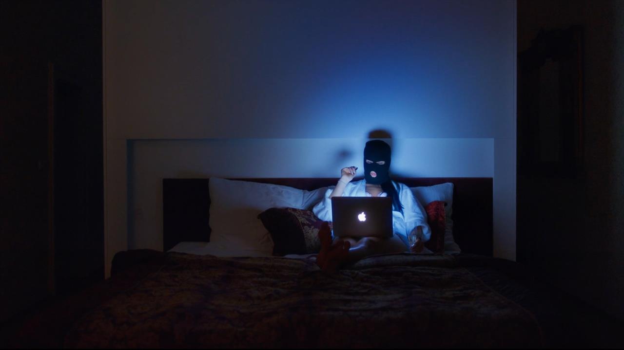 Woman in balaclava sits on a bed with a laptop