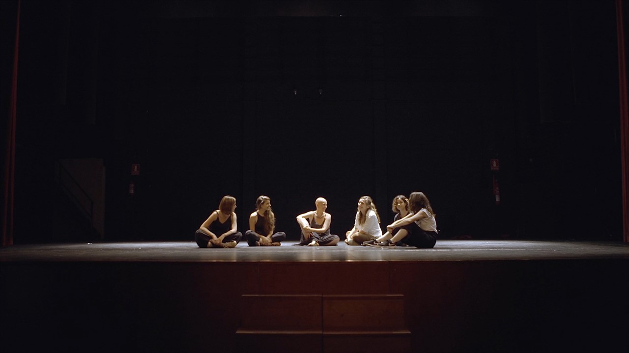 A group of six women sit on an empty stage