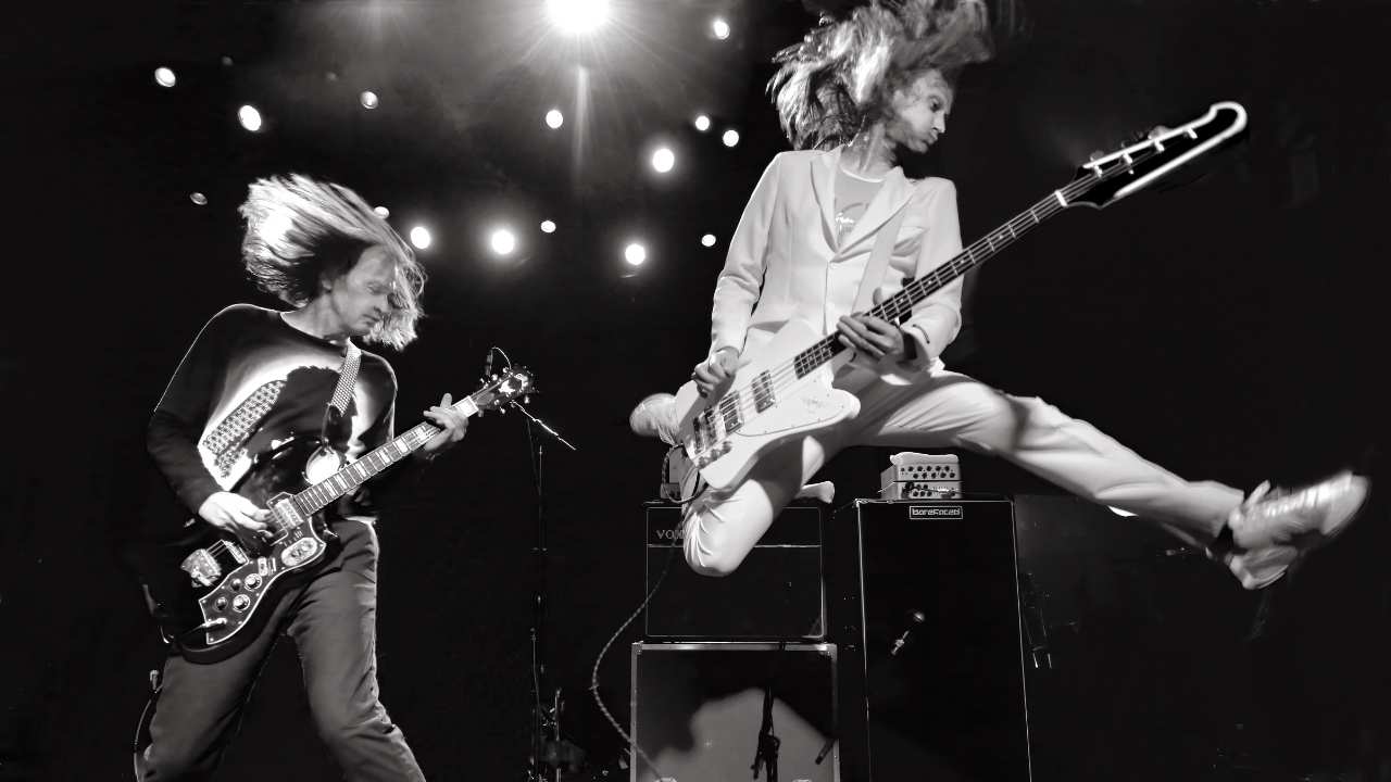 two men with long hair jumping with guitars