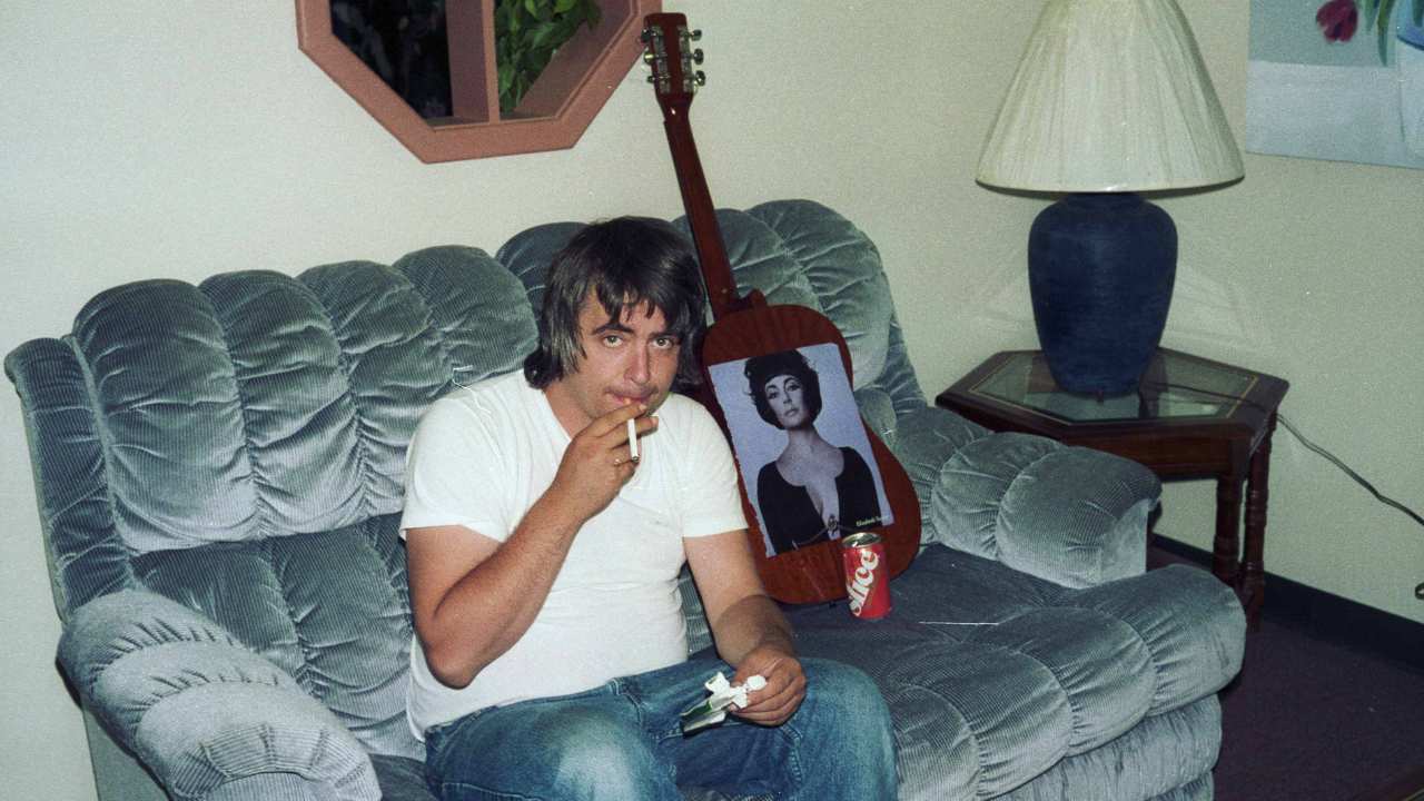 Daniel Johnston smoking on a couch by a guitar