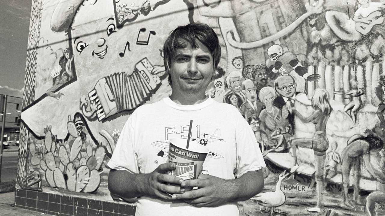 Daniel Johnston holding a cup in front of a mural