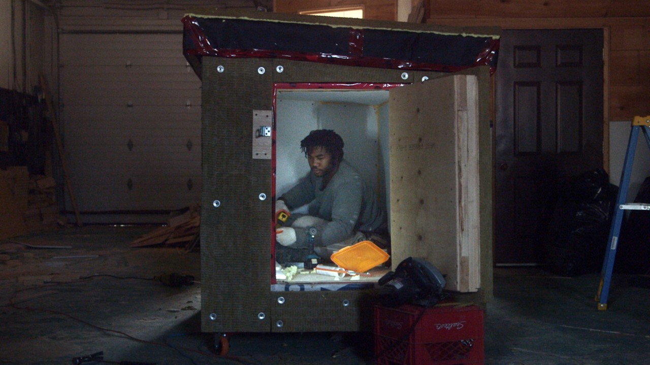 A man sits in a small wooden box with a door on wh